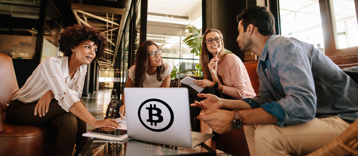 Four professionals sitting around a laptop with a large Bitcoin symbol on the back